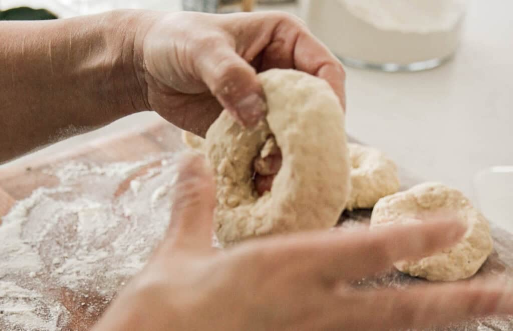 Close-up of hands shaping dough into a ring on a floured wooden surface. Several other dough pieces are visible in the background, with a bowl of flour nearby, suggesting a baking preparation scene.